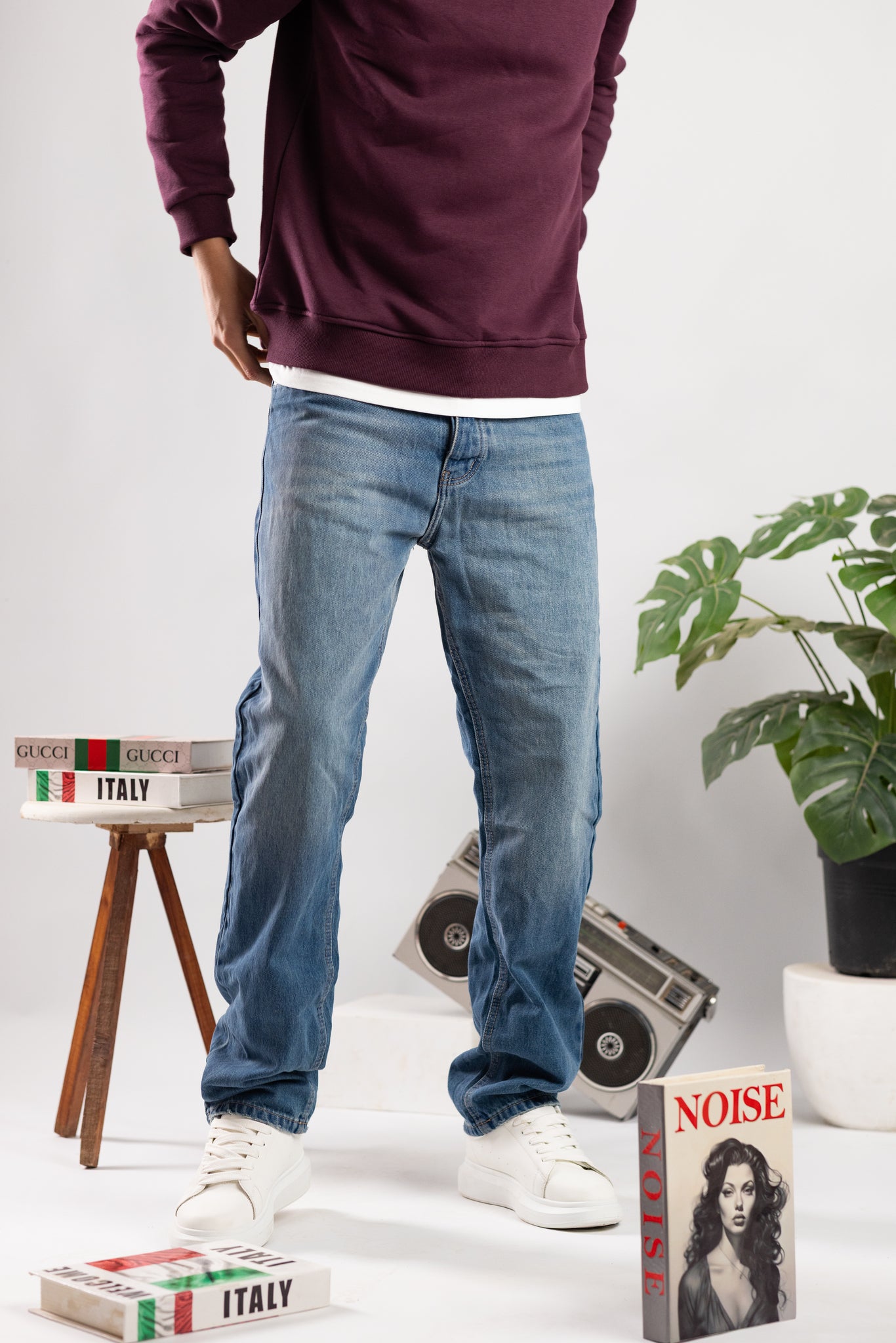 Person wearing a maroon hoodie, blue jeans, and white sneakers standing in a room with books and a plant.